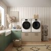 Moderne wasruimte met een wasmachine en droger in de Wasmachine Kast Oslo - Perle Taupe, groene en taupe kasten, decoratieve aanrechtbladen, een gevlochten mand, wasbenodigdheden op een karretje en een zonnig raam.