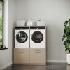 Een wasruimte met de Wasmachine Kast Oslo - Perle Taupe, waarin de wasmachine en de droger naast elkaar staan, met daarboven netjes opgeruimde manden en wasbenodigdheden, een grote groene plant aan de rechterkant en een raam aan de linkerkant dat uitzicht biedt op het weelderige groen buiten.