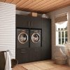 Moderne wasruimte met twee wasmachines met voorlader in donkere kasten, een raam met beige rolgordijn, witte lambrisering en een houten plafond. Handdoeken en wasmanden zijn netjes gerangschikt.