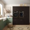 Moderne wasruimte met groene onderkasten, een wit aanrechtblad en gestapelde donkerhouten kasten met daarin een wasmachine en droger met voorlader. Een mand, opgerolde handdoeken en decoratie geven de ruimte warmte. Licht schijnt door een groot raam.