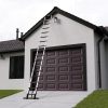 De RMBO® Telescopische ladder 5,1m is tegen het dak geplaatst boven de donkere garagedeur van een modern huis en reikt tot aan een lamp vlakbij de top op een hellende oprit.