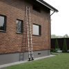 Een RMBO® Telescopische ladder 5,1m leunt tegen het bovenste raam van een bakstenen huis met meerdere ramen, omgeven door een tuin met groen gras, struiken en verschillende kleine bomen.