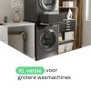 Moderne wasruimte met een gestapelde zwarte wasmachine en droger, waarbij gebruik is gemaakt van het Tussenstuk wasmachine & droger XL Zwart – Universeel, opbergplanken met manden en toiletartikelen, een stoel en een decoratieve vaas. De Nederlandse tekst luidt: XL-versie voor grotere wasmachines.